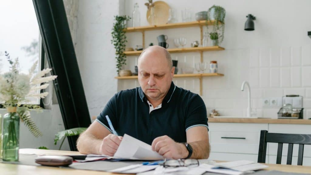 A bald man in a polo shirt sits at a kitchen table reviewing and signing paperwork.