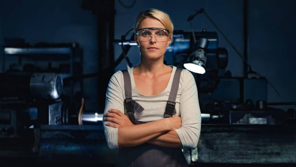 A woman standing with arms crossed indoors in a metal workshop looking at the camera.