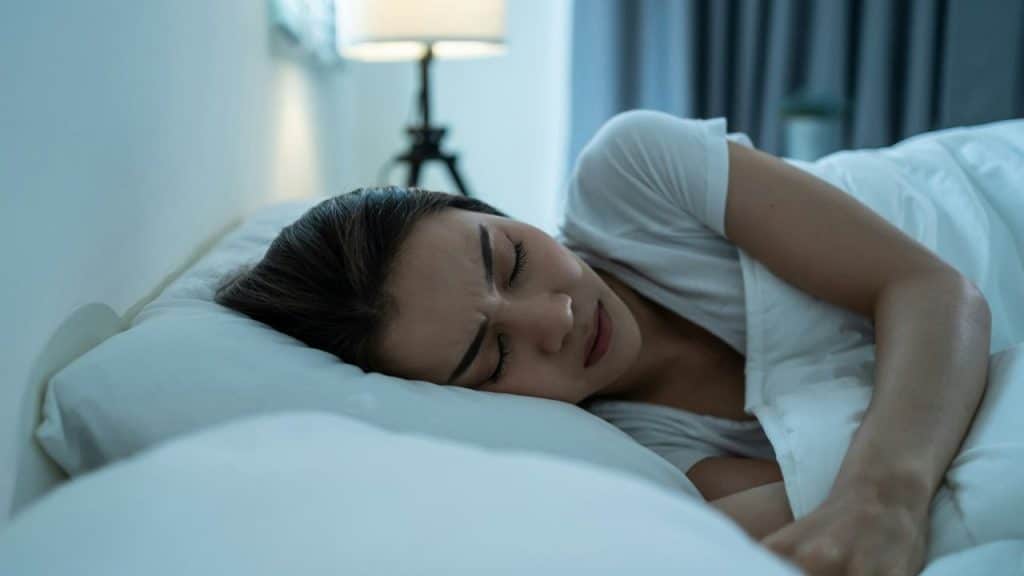 A woman lies in bed with her eyes closed and face somewhat contorted, as if in discomfort.