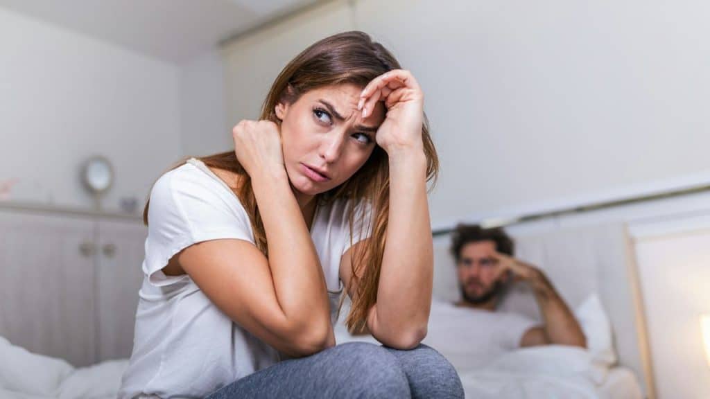 A woman sits on a bed looking sideways with her hand raised to her forehead.