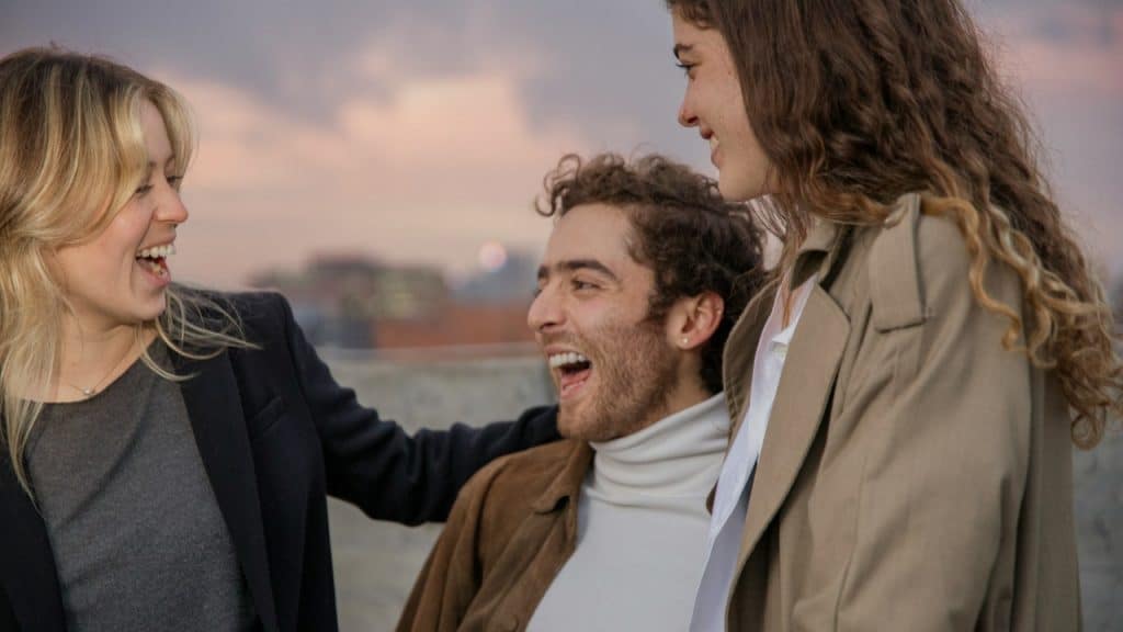 Three people stand outdoors on a rooftop, laughing together with a city skyline in the background.