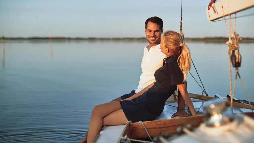 A couple sitting on a sailboat, smiling at each other on calm water.