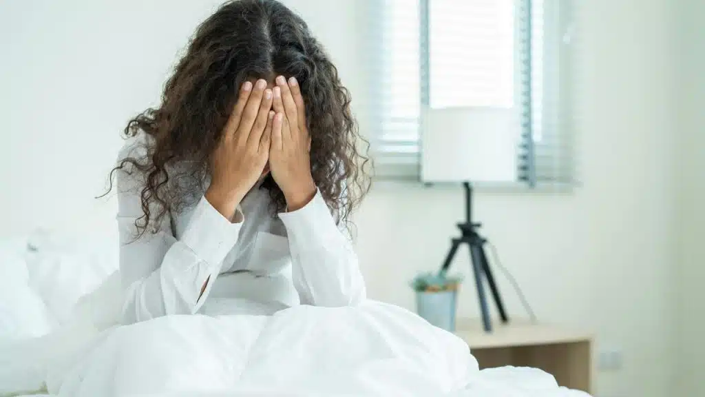 A woman sits on a bed with her face covered by her hands.