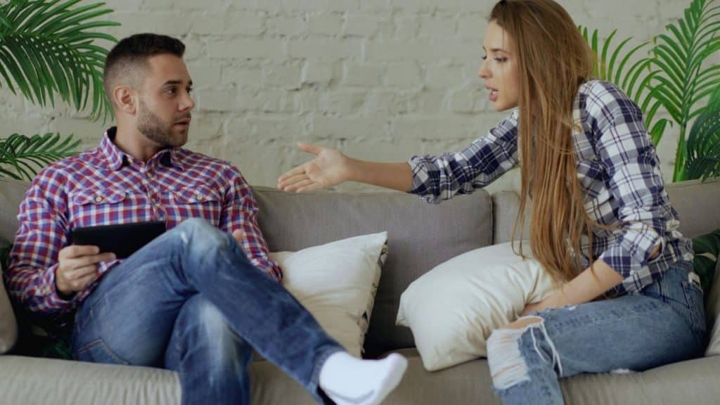 A woman in a plaid shirt gestures toward a man sitting on a sofa.