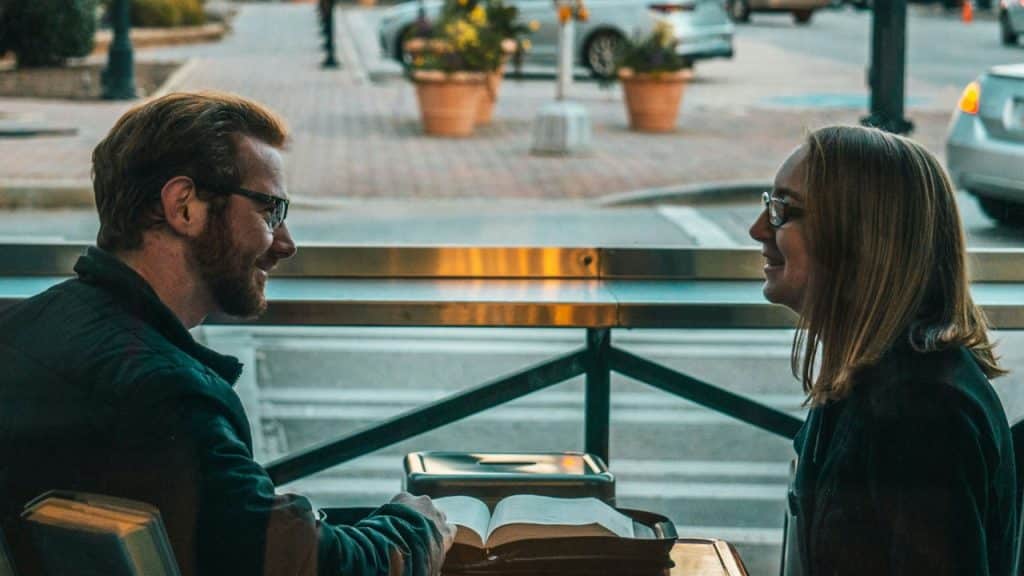 A man and woman with glasses face each other across a table with an open book.