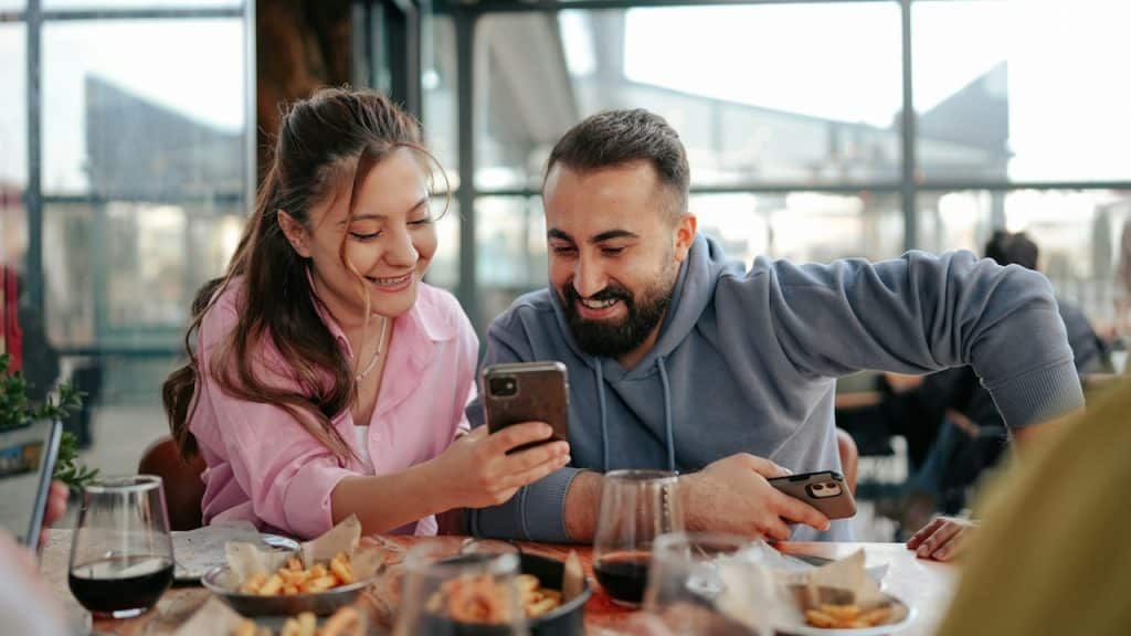 A smiling woman is showing something to a smiling bearded man on her phone as they sit at a table.