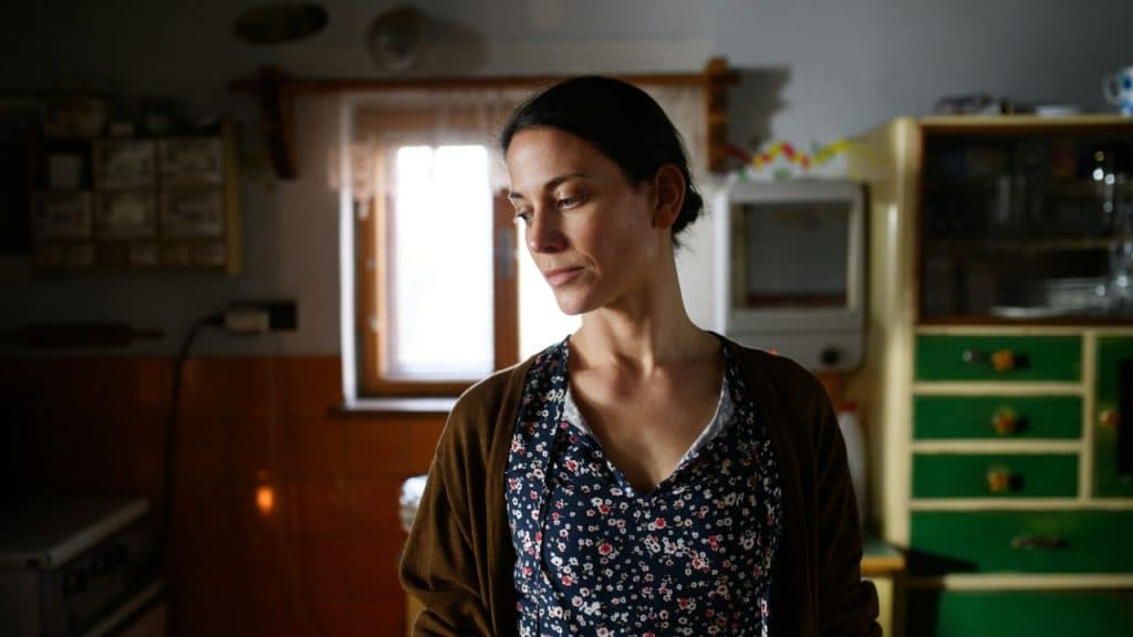 A woman with dark hair tied back looks downward with a somber expression in a kitchen.