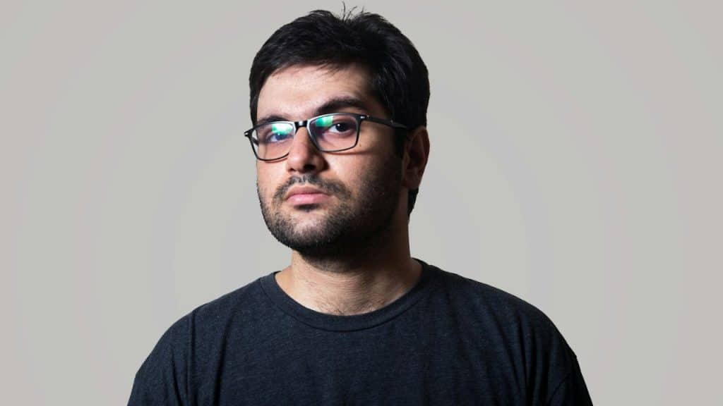 A solemn man with glasses and a beard stares at the camera.