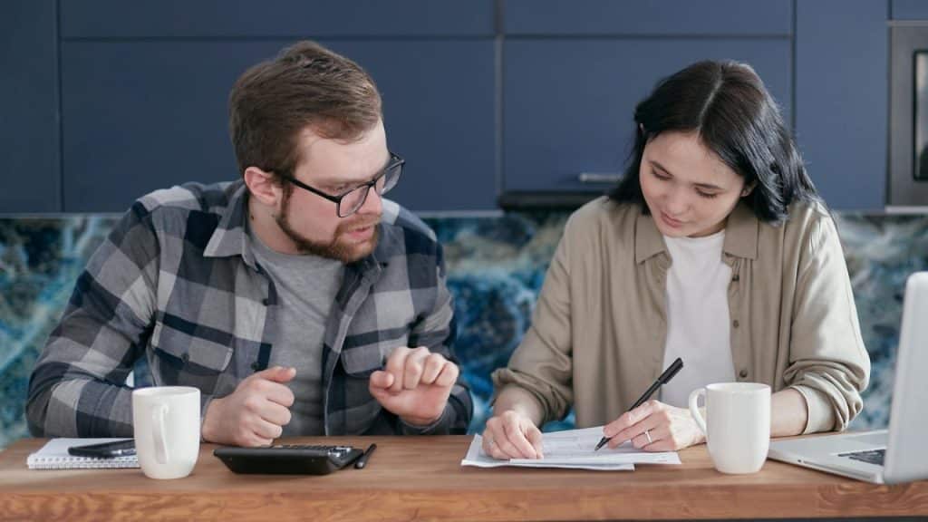 A man and woman reviewing paperwork together at a table with a calculator and coffee mugs.