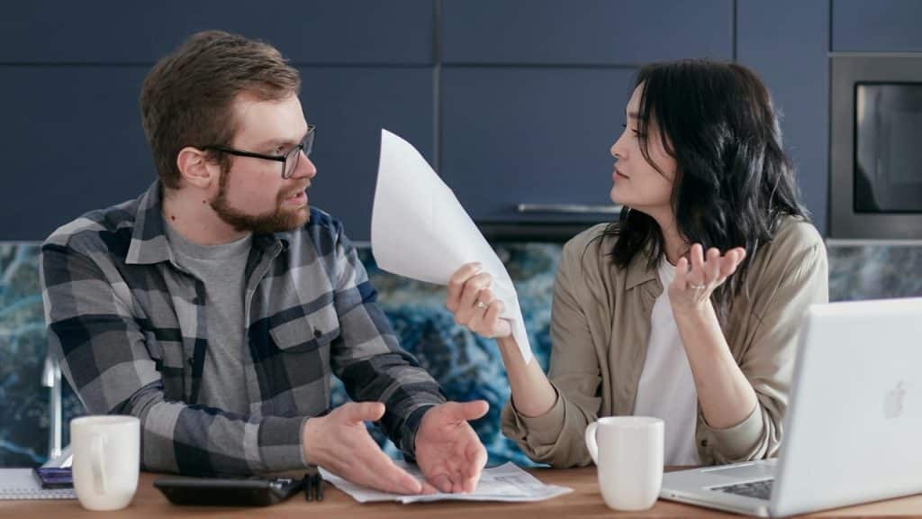 A man and woman sit at a table with papers, a laptop, and two mugs.