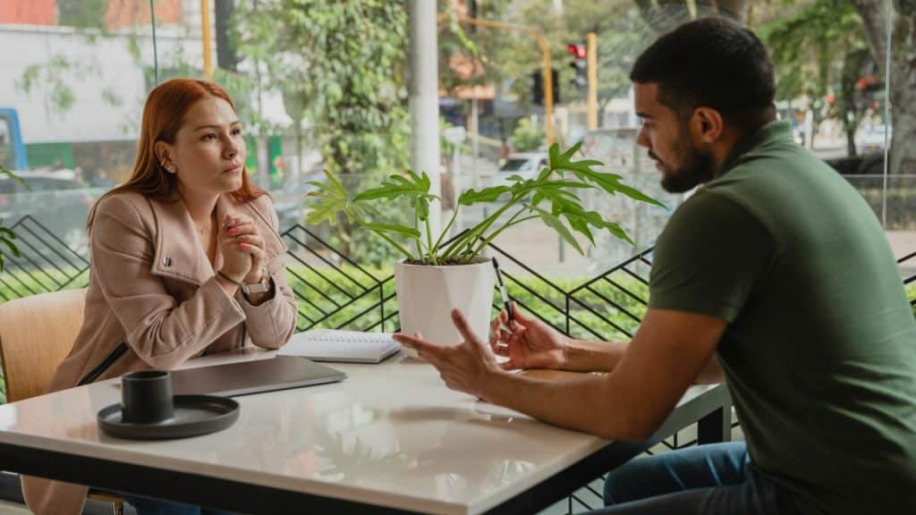 A woman listens intently as a man gestures while speaking to her across a cafe table.
