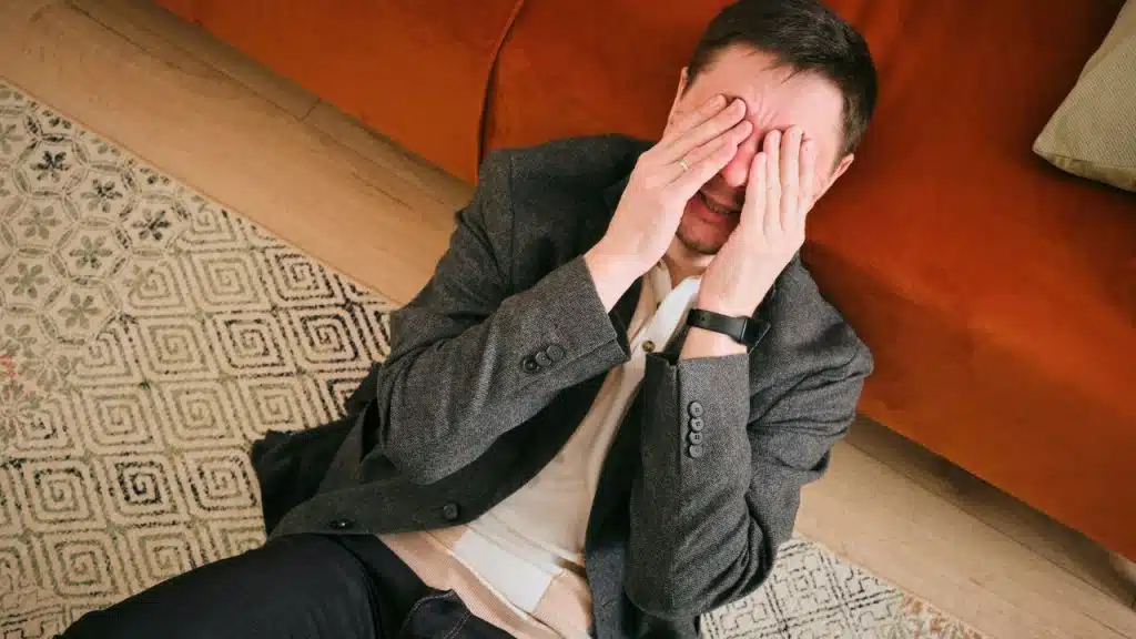 A man in a grey jacket sits on the floor covering his eyes with his hands.