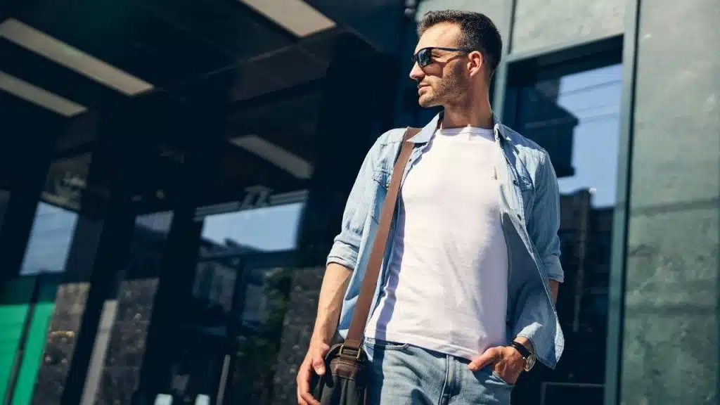 A man in a denim shirt and sunglasses stands outside a modern glass building.