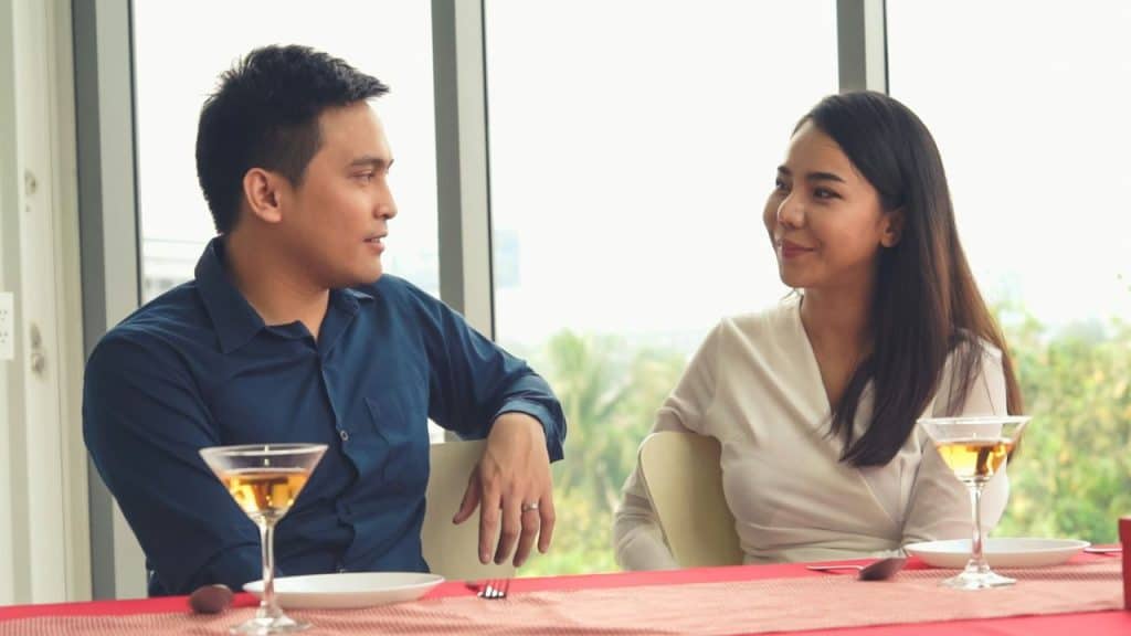 A man and a woman sit at a table with drinks, looking at each other.