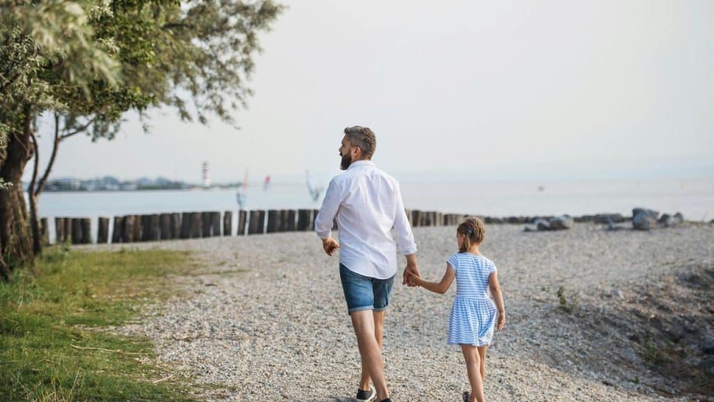 A father and daughter walking hand in hand on a pebble beach.