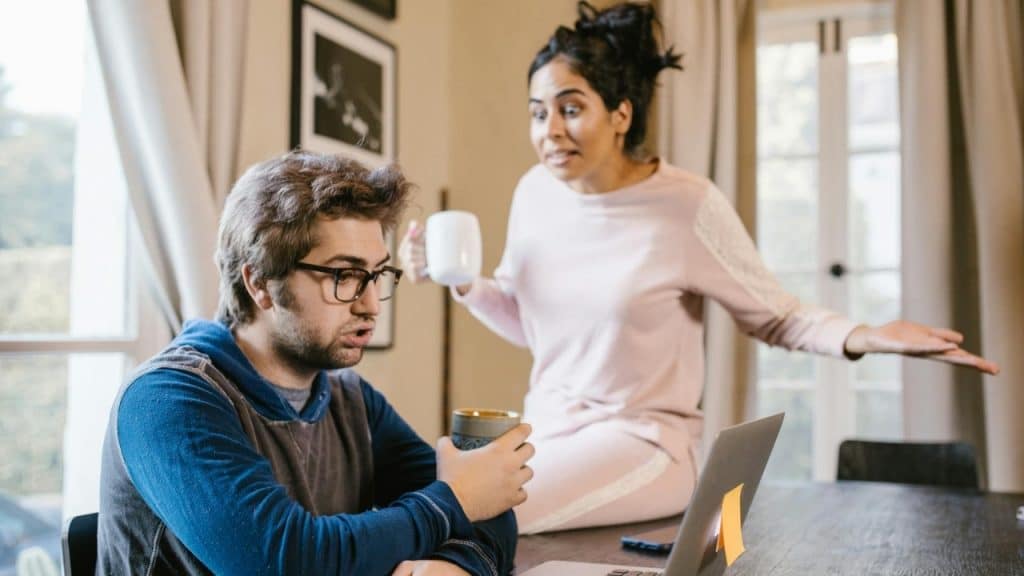 A woman stands and gestures with an open hand toward a man sitting at a laptop.