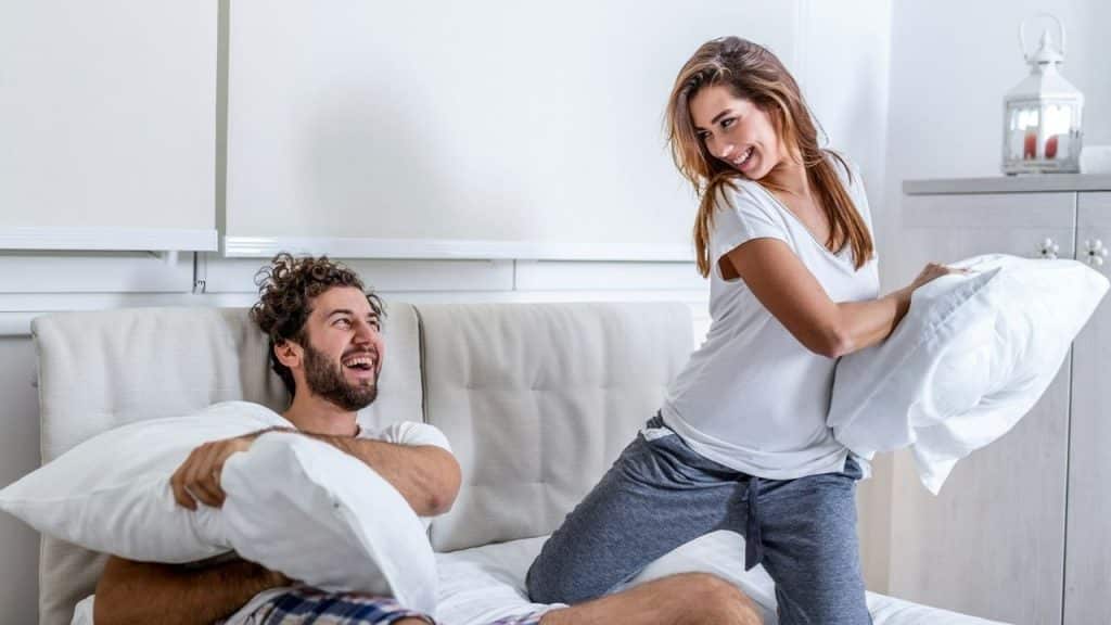 A man and woman laugh while engaging in a playful pillow fight on a bed.
