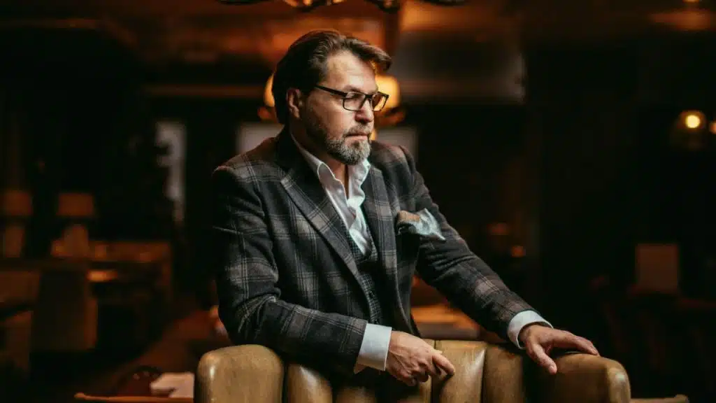 A man in a plaid suit jacket leans on a leather chair in a dimly lit room.