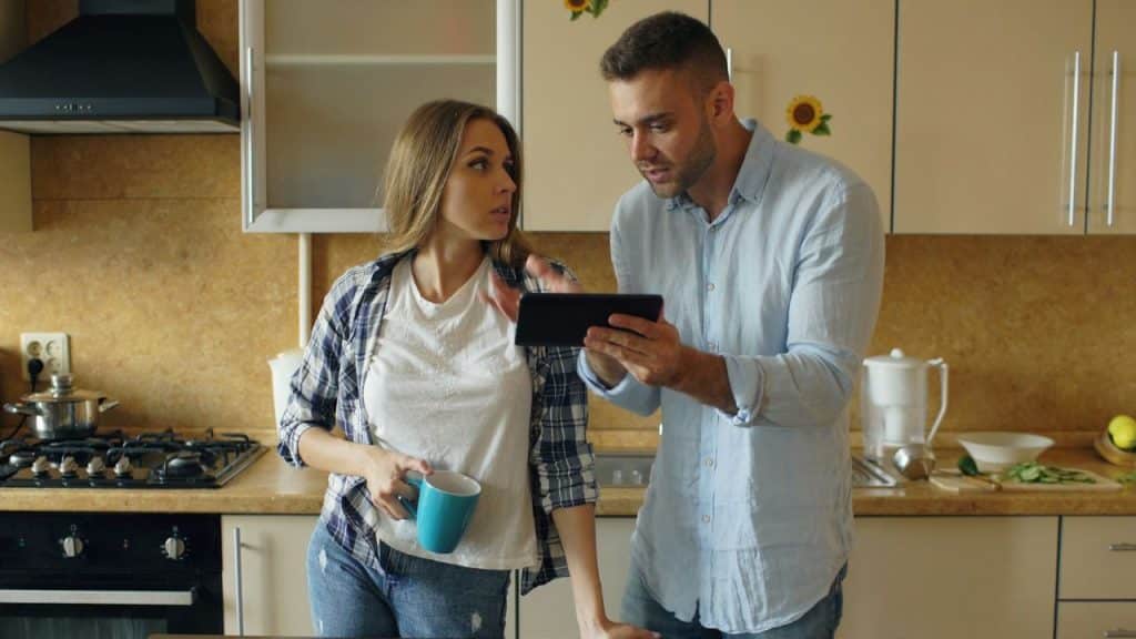 A man holds a tablet and speaks to a woman holding a blue mug in a kitchen.