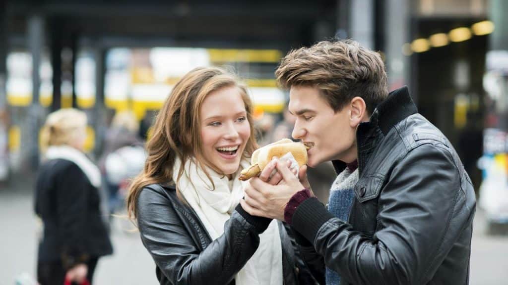 A young couple laughing while sharing a sandwich outdoors.