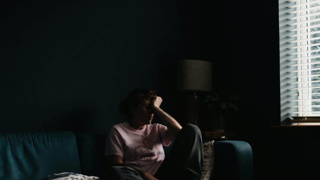 A person sitting on a couch in a dark room with one hand on their forehead near a window.