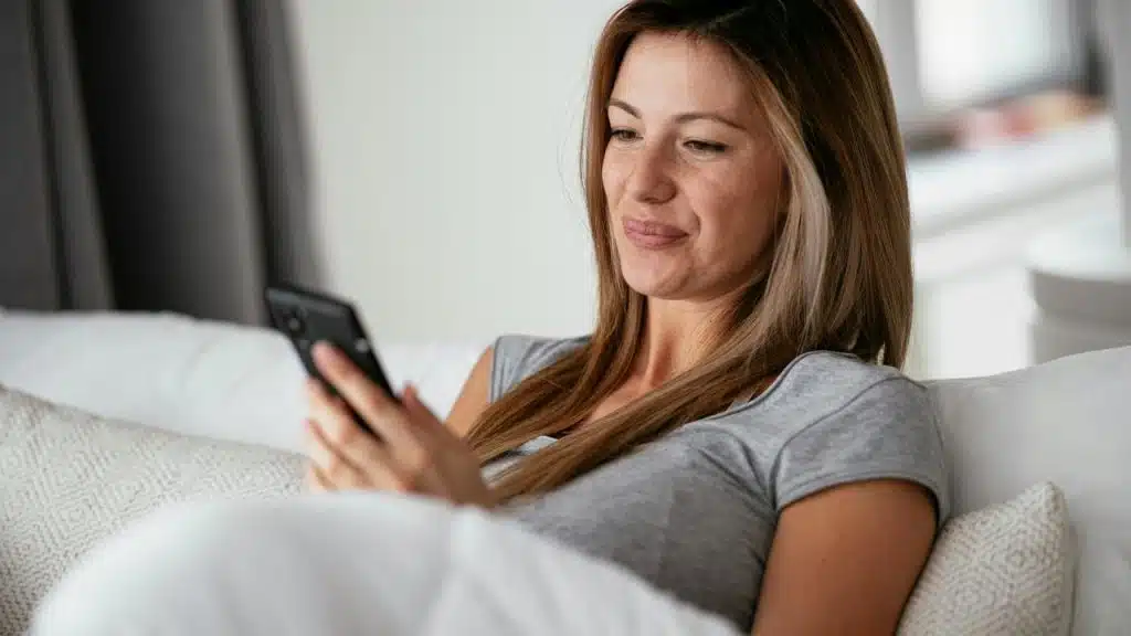A woman smiling while using a smartphone on a couch.