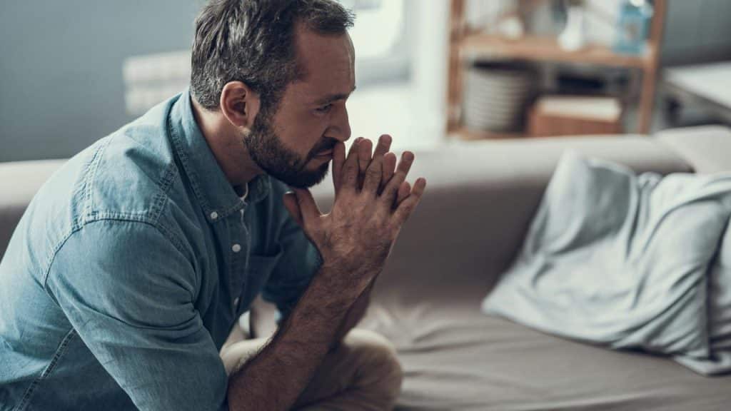 A man sitting on a couch with his hands clasped in thought.