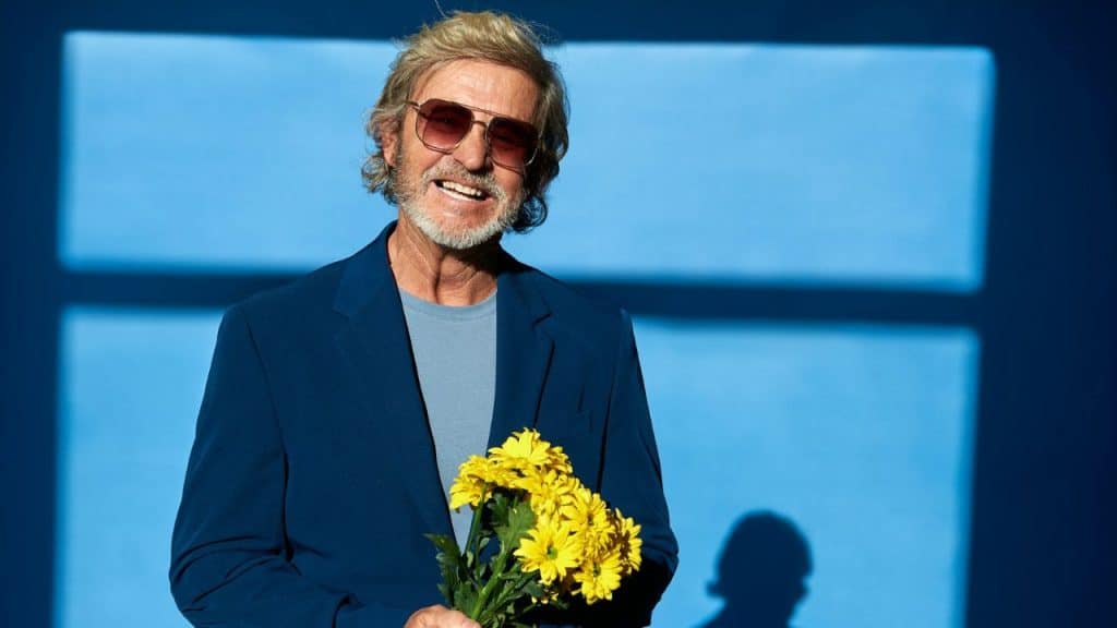 A smiling older man with blonde hair wears sunglasses and a blue suit holding yellow flowers.