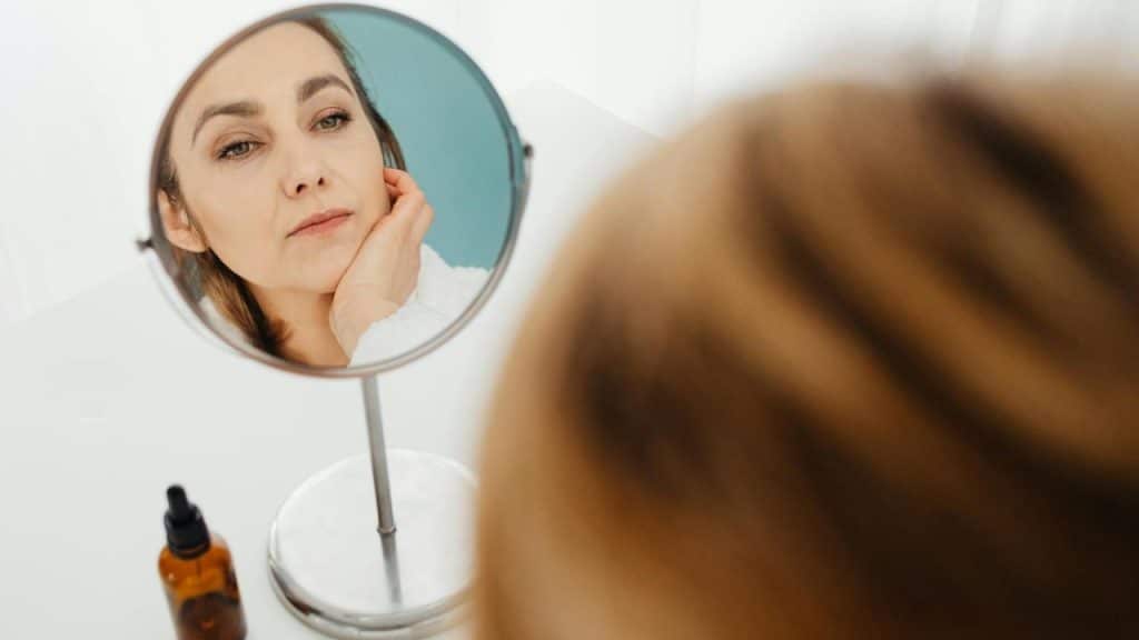 A woman looks into a circular vanity mirror while holding her hand against her chin.