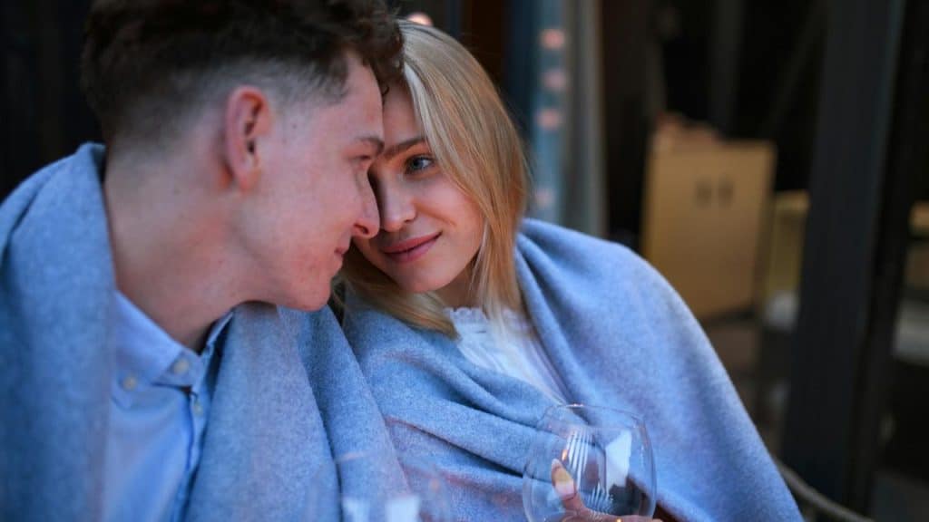 A couple wrapped in a blanket sharing an intimate moment while holding wine glasses outdoors.