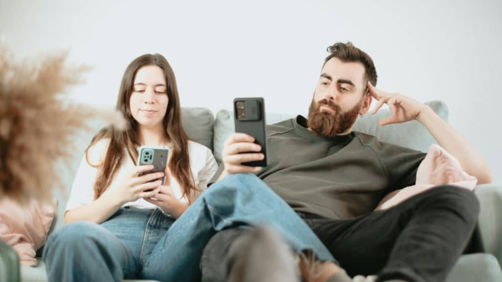 A man and woman sit on a sofa together while both looking down at their own smartphones.