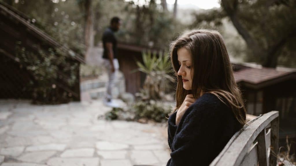 A woman sitting sadly on an outdoor bench with a man standing in the blurry background.