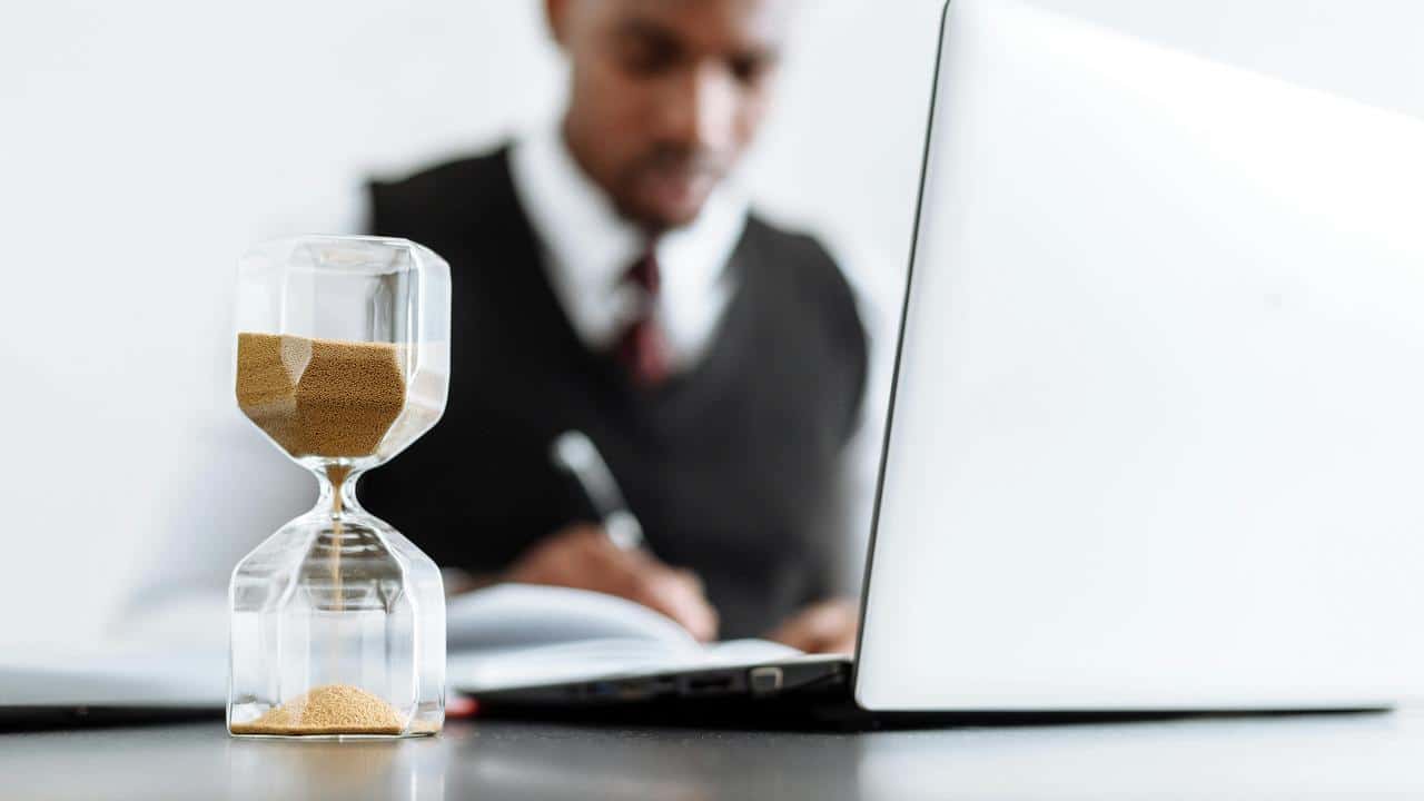 An hourglass on a desk next to a man writing.