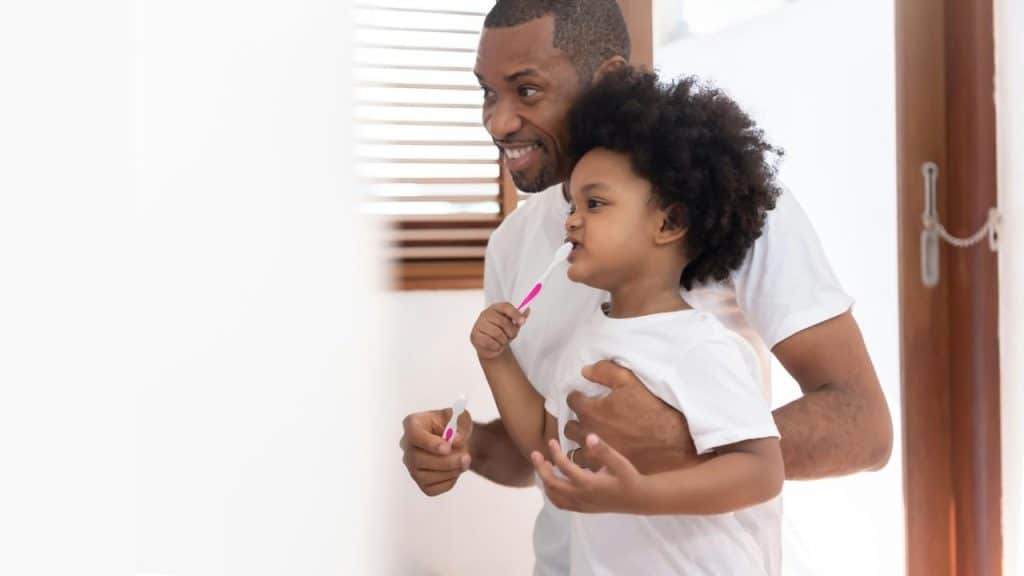 A father helping his young child brush their teeth in a bright bathroom.