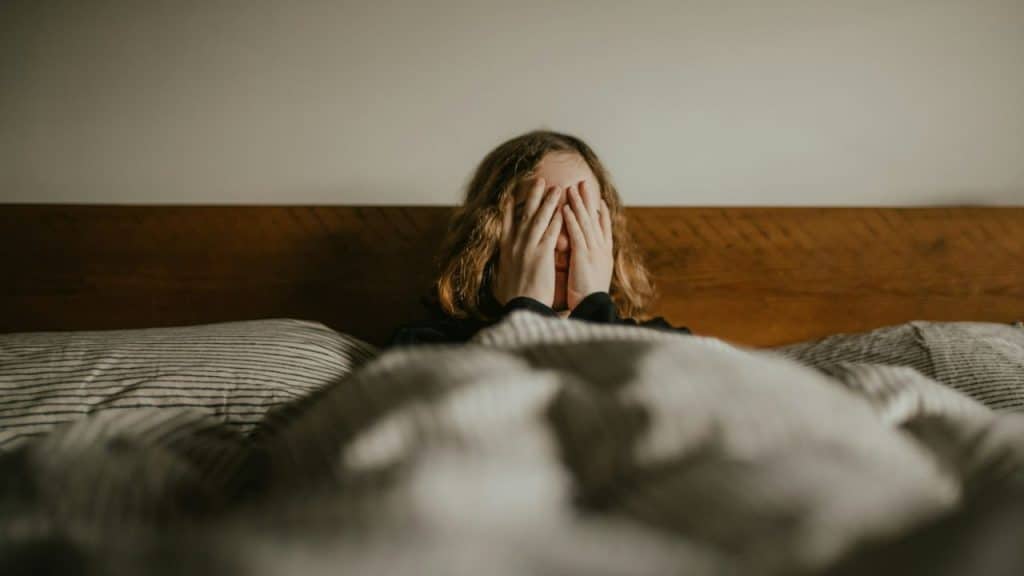 A person in a bed covers their face with both hands while lying under blankets.