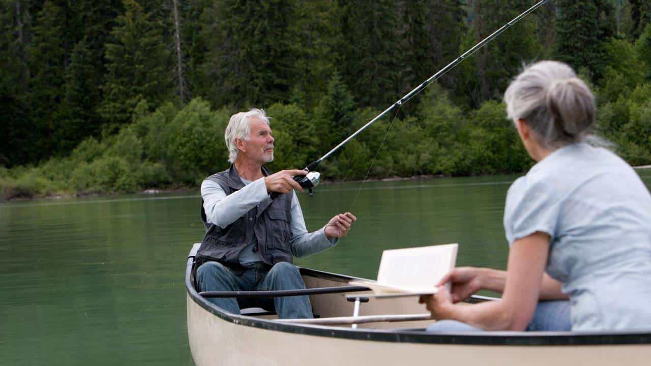 An elderly man fishing from a canoe as a woman reads beside him.