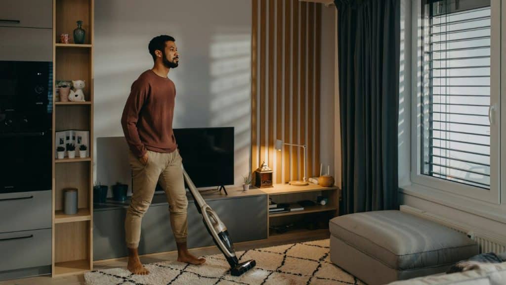 A man stands on a rug holding a vacuum while looking out a nearby window.