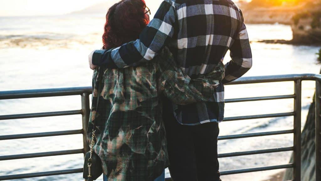A couple standing arm in arm by a railing, looking out at the ocean during sunset.