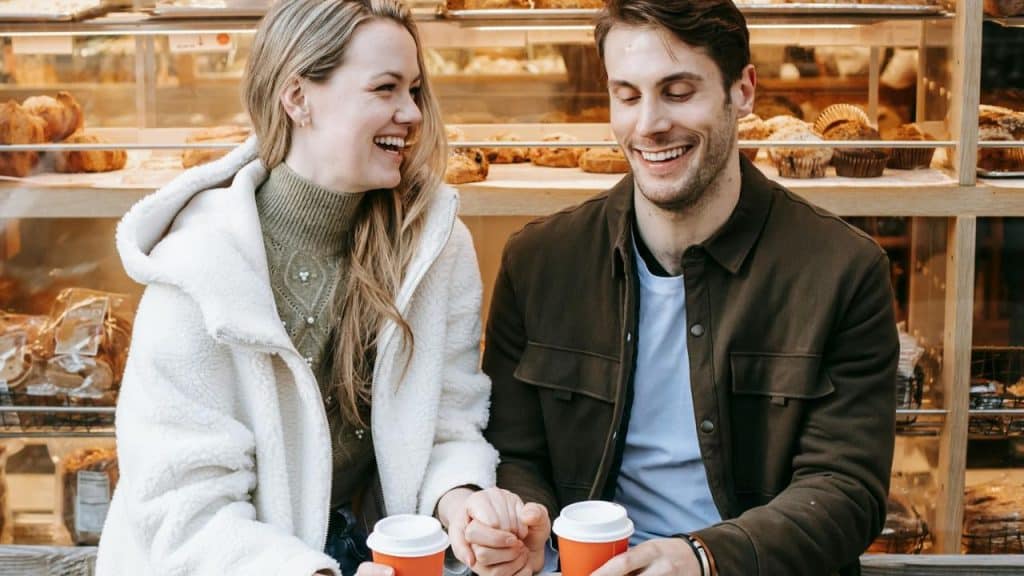 A laughing man and woman sit in front of a bakery display holding coffee cups.