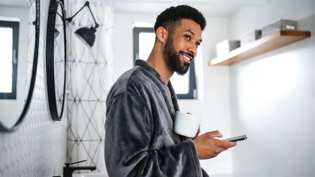 A smiling man in a grey robe holds a white mug and a smartphone indoors.