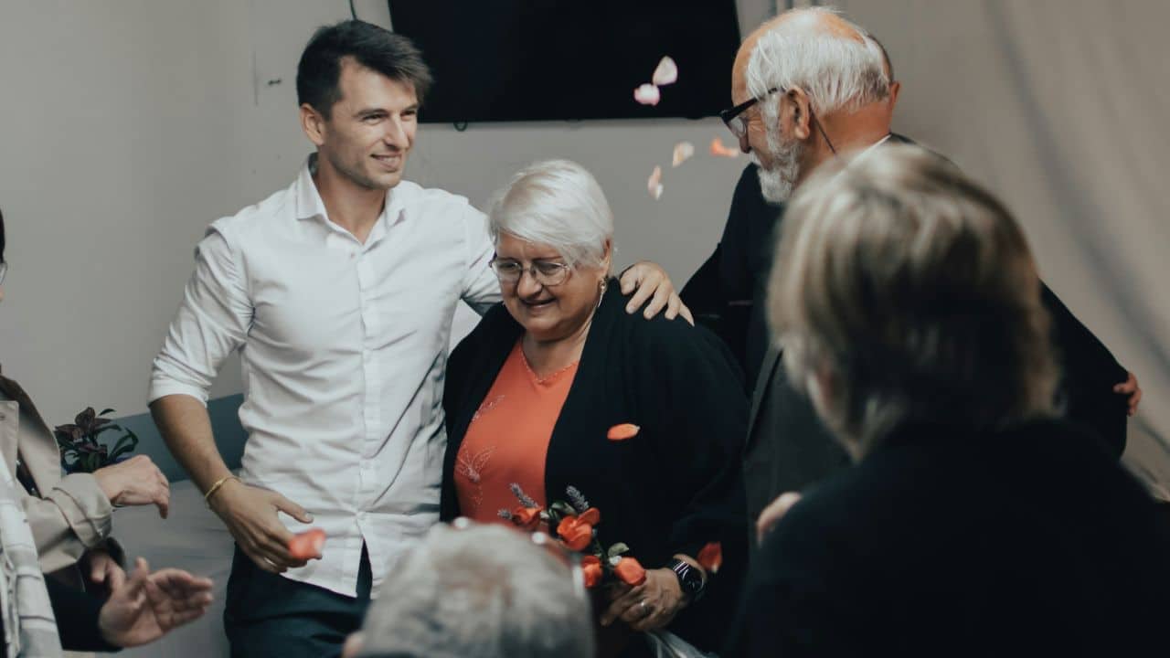 A younger man smiles while standing next to an older woman as flower petals fall.