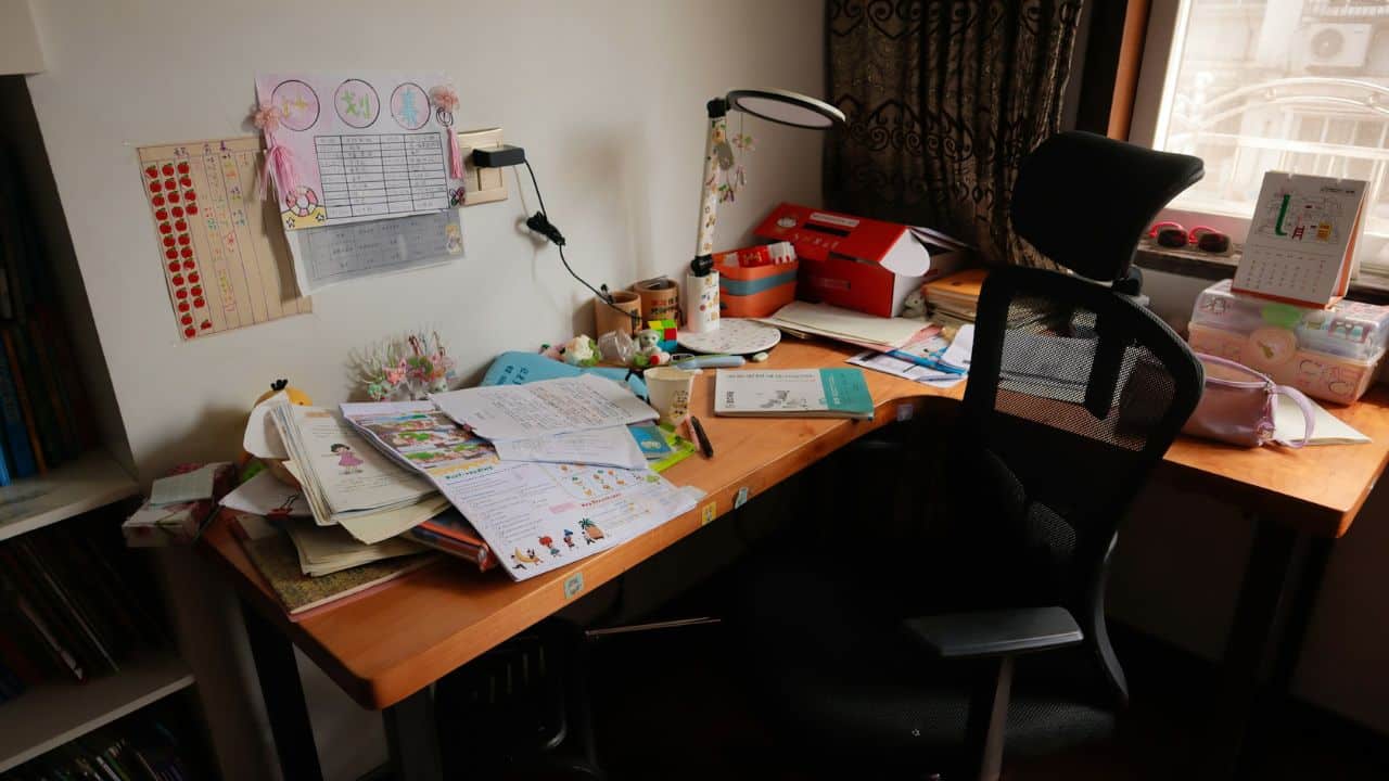 A wooden desk is cluttered with various papers, books, a lamp, and a black office chair.