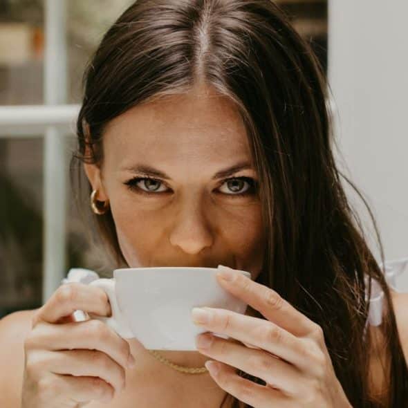 A woman with long brown hair looks forward while drinking from a white ceramic cup.
