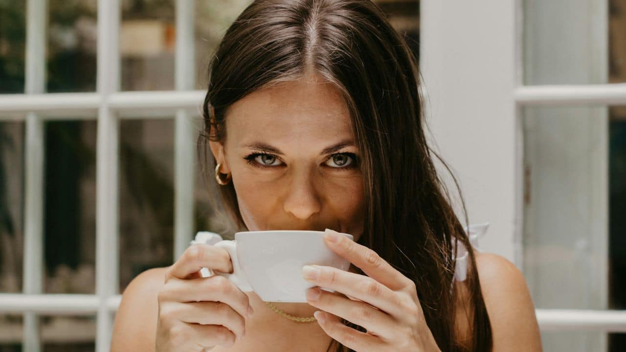 A woman with long brown hair looks forward while drinking from a white ceramic cup.