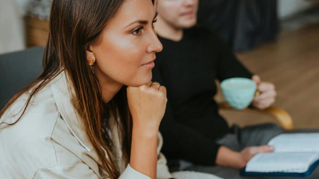 A woman listening thoughtfully while sitting beside someone holding a book and a cup.