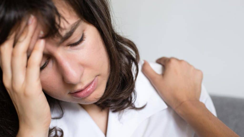 A woman with her eyes closed touching her forehead while another hand rests on her shoulder.