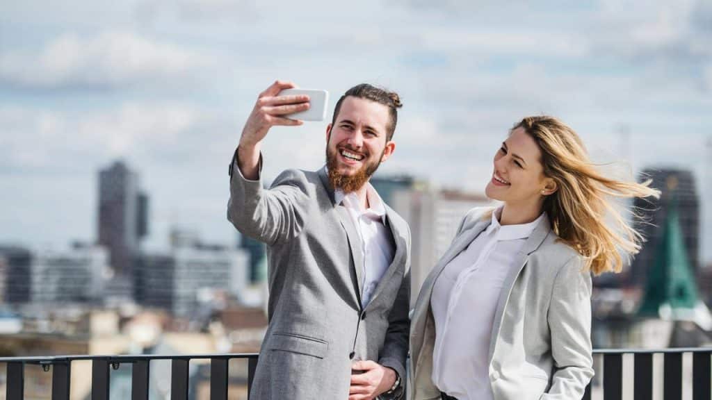 A smiling man and woman taking a selfie on a rooftop with a city skyline behind them.