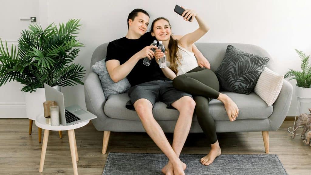 A couple relaxing on a couch while taking a selfie and holding water bottles.