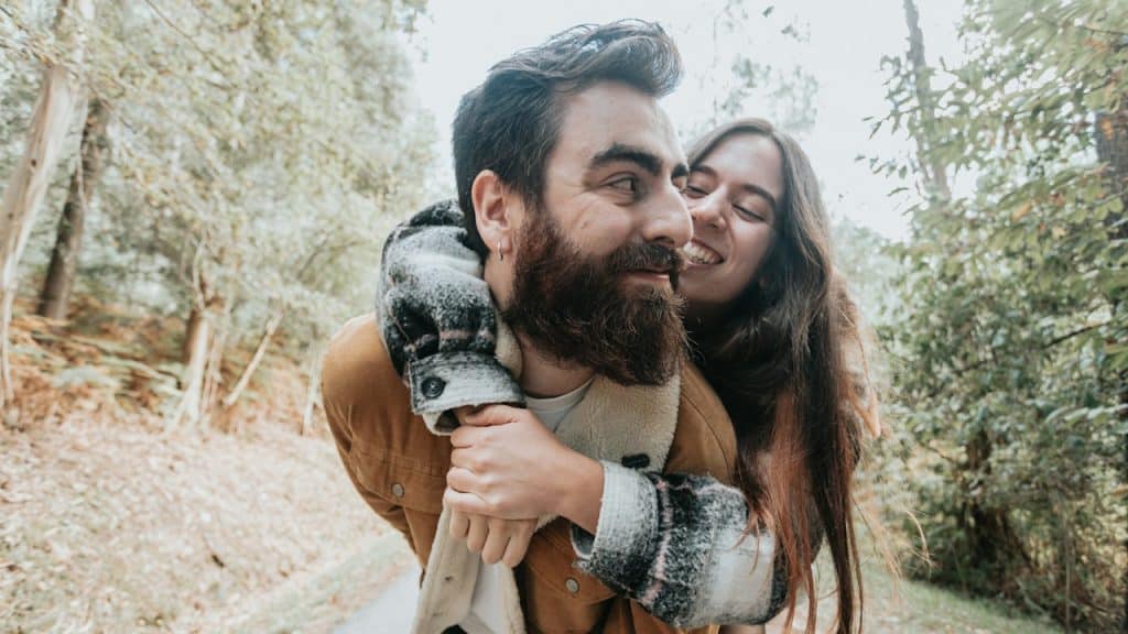 A woman riding on the back of his husband while they are walking in the forest.