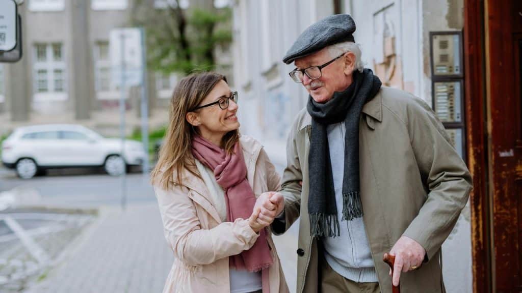 A woman and an older man in a flat cap walk outside while holding hands together.